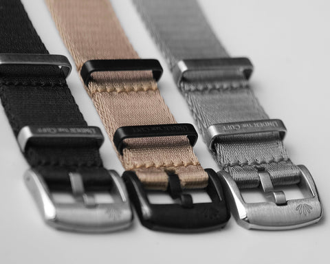 Selection of Fineweave NATO straps in black, tan, and grey, each featuring a sleek buckle, meticulously aligned on a white surface - Under the Cuff's refined Fineweave NATO Collection.